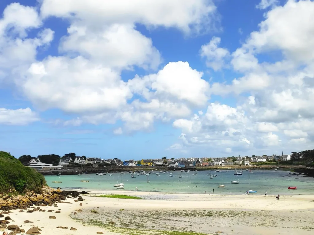 Beniget près de portsall maison de vacances Cette photo capture une plage pittoresque près de Portsall. Le sable est fin et clair, bordé de rochers épars. Plusieurs bateaux sont ancrés dans l'eau calme, transparente, reflétant un ciel bleu parsemé de nuages blancs volumineux. Au fond, on aperçoit le village de Portsall, avec ses maisons traditionnelles bretonnes alignées le long du rivage. Ce lieu tranquille et charmant est typique des maisons de vacances recherchées dans cette région pour leur vue imprenable et leur accès direct à la mer.