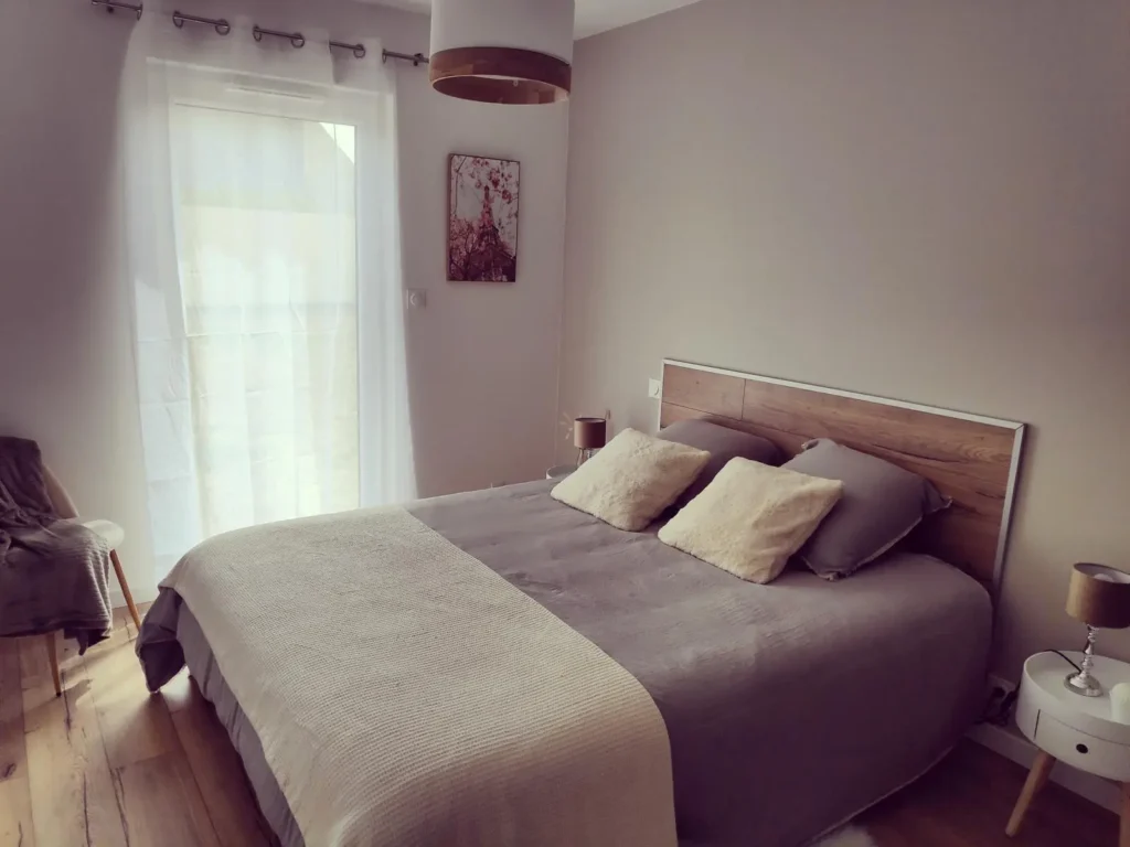 Chambre maison de vacance portsall Cette image montre une chambre à coucher dans une maison de vacances à Portsall. La pièce est décorée dans des tons neutres, avec un grand lit double au centre, doté d'une tête de lit en bois et recouvert d'une literie grise et de coussins assortis. À côté du lit, une petite table de nuit blanche avec une lampe. Sur le mur, un tableau avec des motifs rouges ajoute une touche de couleur. Une grande fenêtre drapée d'un rideau blanc laisse entrer la lumière naturelle, créant une ambiance douce et reposante.