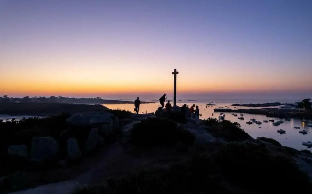 Mégalithes du Guilliguy L'image montre un paysage au crépuscule avec des mégalithes du Guilliguy au premier plan, formant un chemin qui mène à une croix sur un promontoire où quelques personnes se sont rassemblées. Le fond offre une vue sur une baie calme avec des petits bateaux et la silhouette d'une maison de location à Portsall. Le ciel se pare de teintes violettes et orangées, ajoutant une atmosphère paisible à la scène.