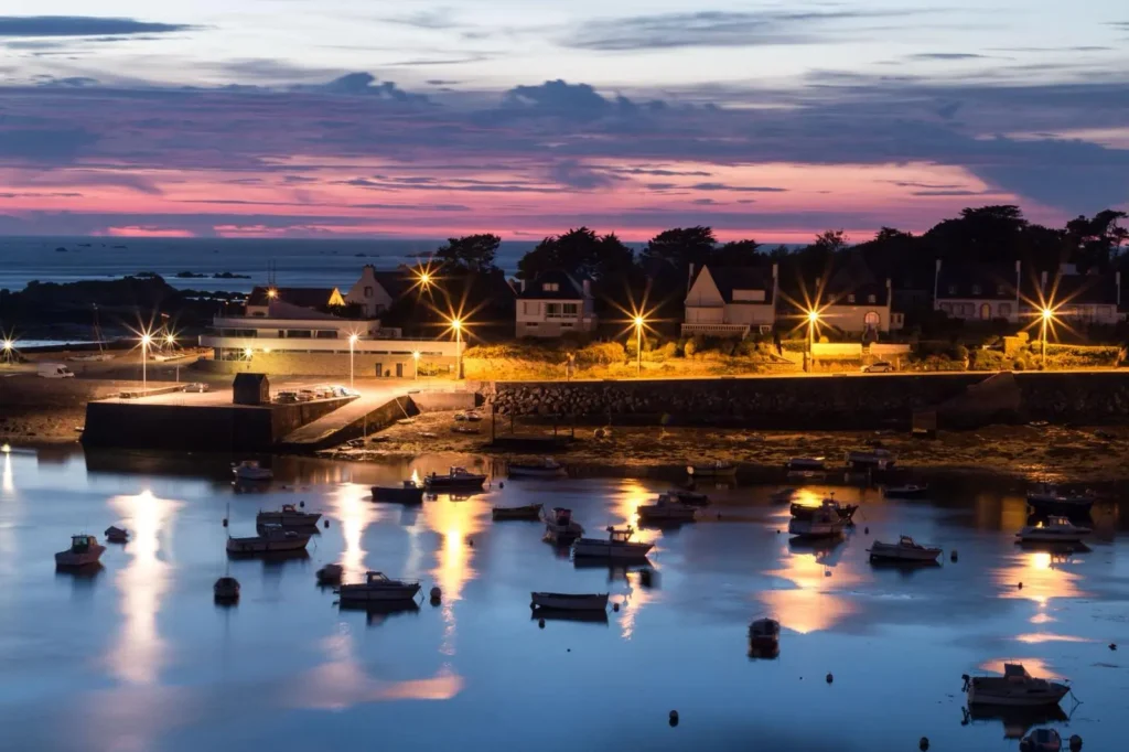 Port de Portsall Cette image montre le port de Portsall au crépuscule, avec une vue magnifique sur des bateaux amarrés dans des eaux calmes, réfléchissant les lumières jaunes des lampadaires environnants. Le ciel est teinté de nuances de rose et de violet au-dessus d'une ligne d'horizon parsemée d'îlots rocheux. À l'arrière-plan, des maisons typiques de la région suggèrent des locations de vacances à Portsall, offrant une proximité immédiate avec le port et les commerces locaux éclairés, soulignant la convivialité et l'accessibilité de ce charmant village côtier.
