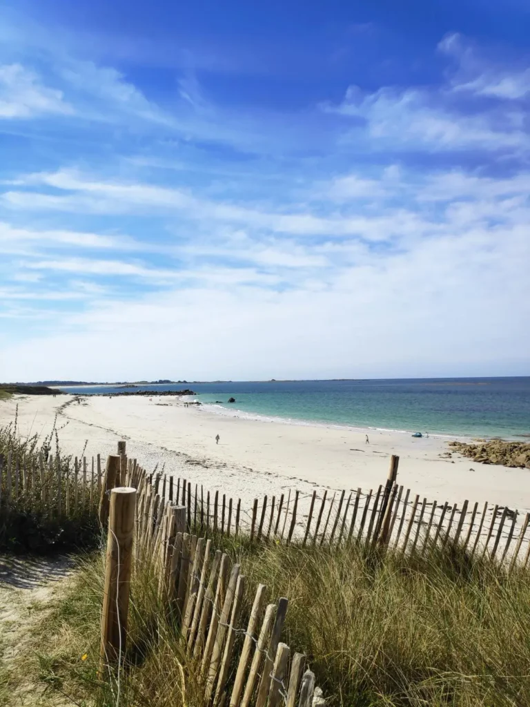 Sables fin portsall Cette image illustre parfaitement le charme des plages près de Portsall, idéales pour une location de maison de vacances. La plage de sable fin s'étend majestueusement sous un ciel bleu clair, parsemé de nuages légers. Une clôture en bois naturel délimite le chemin d'accès à la plage, suggérant un environnement préservé et tranquille. Au loin, quelques personnes profitent du vaste espace, renforçant l'attractivité de ce lieu pour des vacances relaxantes au bord de la mer. C'est un cadre idéal pour ceux qui recherchent la proximité avec la nature sans renoncer aux commodités des commerces proches.