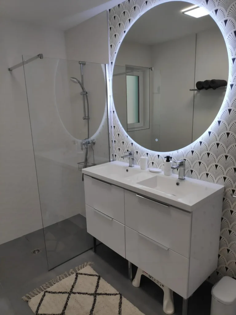 Salle de bain maison de vacance à Portsall Cette image présente une salle de bain élégante d'une maison de vacances à Portsall. Elle est équipée d'une grande douche à l'italienne avec paroi en verre et d'un meuble-lavabo blanc moderne avec un grand tiroir pour le rangement. Un miroir circulaire, entouré d'une lumière LED intégrée, domine le lavabo, reflétant un mur décoré de carreaux à motifs géométriques en relief. Le sol est recouvert de carrelage gris foncé, contrastant avec un tapis blanc et noir à motifs, ajoutant une touche de chaleur et de style à l'espace. Cette salle de bain combine fonctionnalité et design contemporain, créant un environnement relaxant et raffiné.