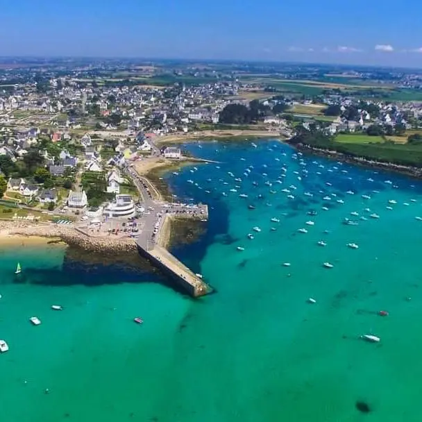 L'image montre une vue aérienne de Portsall, une ville côtière caractérisée par ses eaux cristallines de couleur turquoise. Le port est protégé par une grande digue et rempli de nombreux bateaux de différentes tailles. La ville elle-même est composée de bâtiments dispersés, principalement de petites maisons blanches, entourées de végétation verte. C'est un cadre idyllique et paisible, typique d'une destination de vacances en Bretagne, invitant à la détente et à la découverte marine.